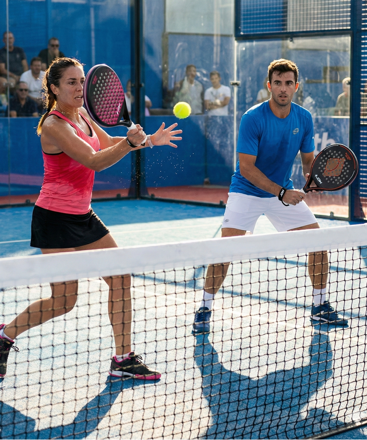 Two people playing padel on a court with spectators in the background
